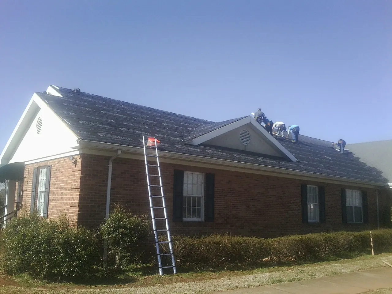 Crew tearing shingles off a brick ranch roof with a ladder set — Emergency Roof Repair work by our crew in Acton